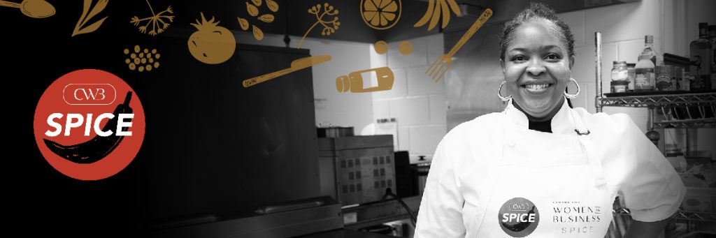 Front profile of a smiling woman wearing an apron and chef jacket standing in an industrial kitchen