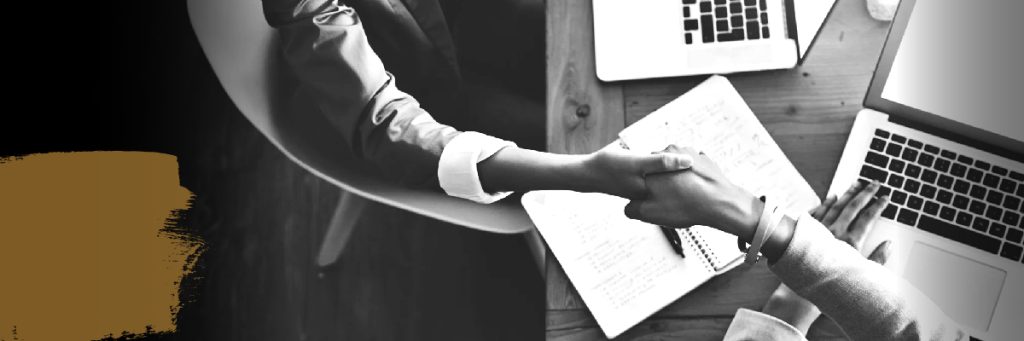 overhead image of two women shaking hands while sitting at a work table with their laptops