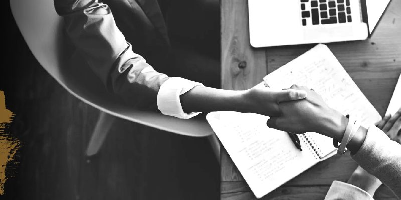 overhead image of two women shaking hands while sitting at a work table with their laptops